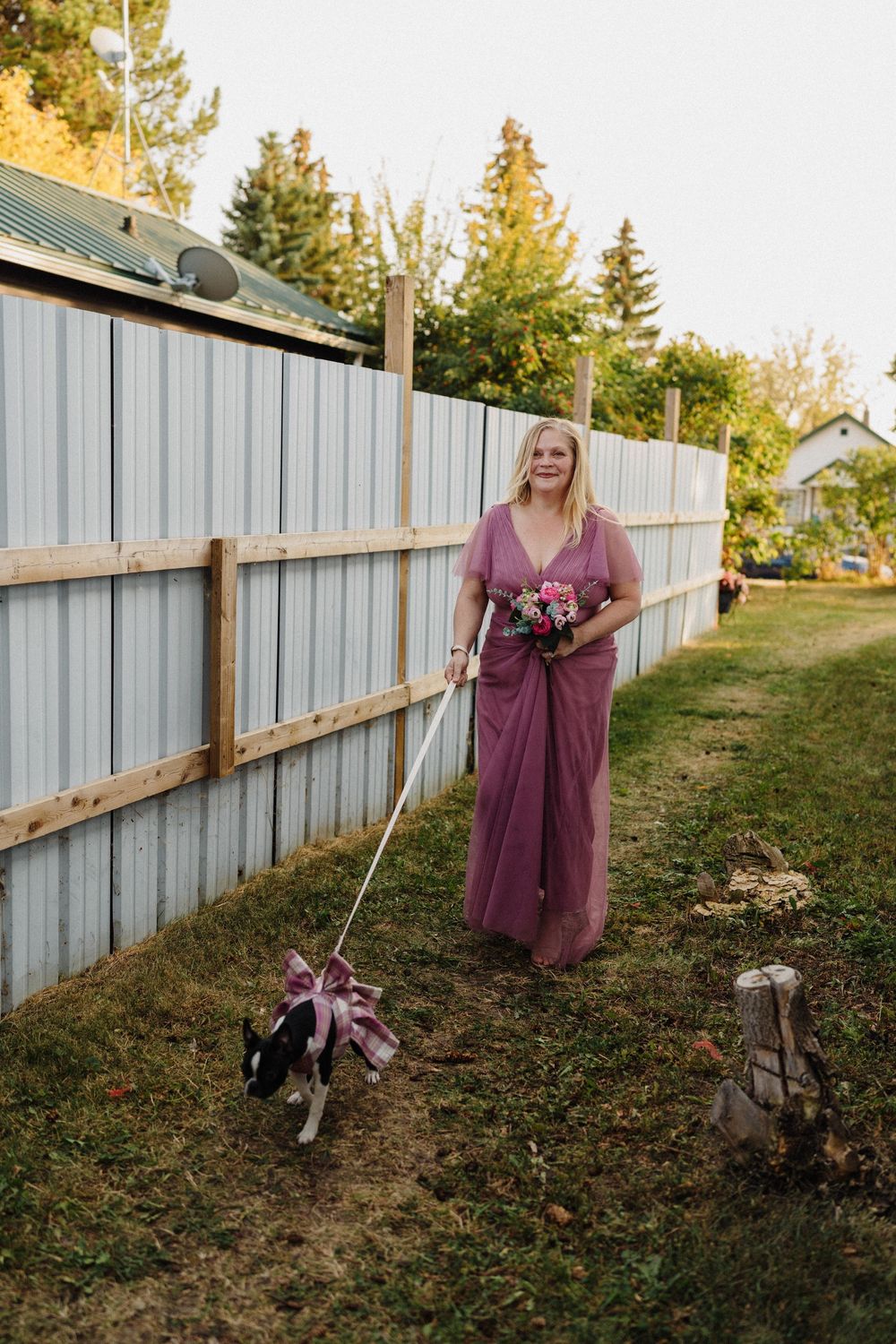 Blonde bridesmaid in dusty rose tulle gown holds floral bouquet while walking a small dog on a leash outdoors.