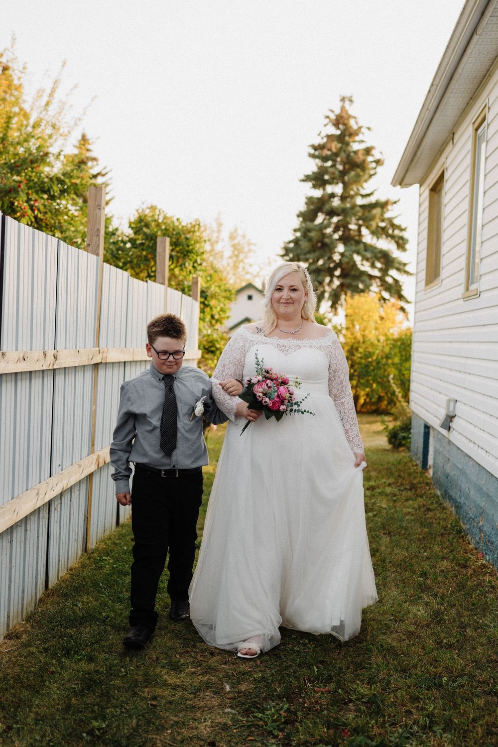 Bride in white lace wedding gown holds pink bouquet while walking with young boy escort in gray suit outdoors.
