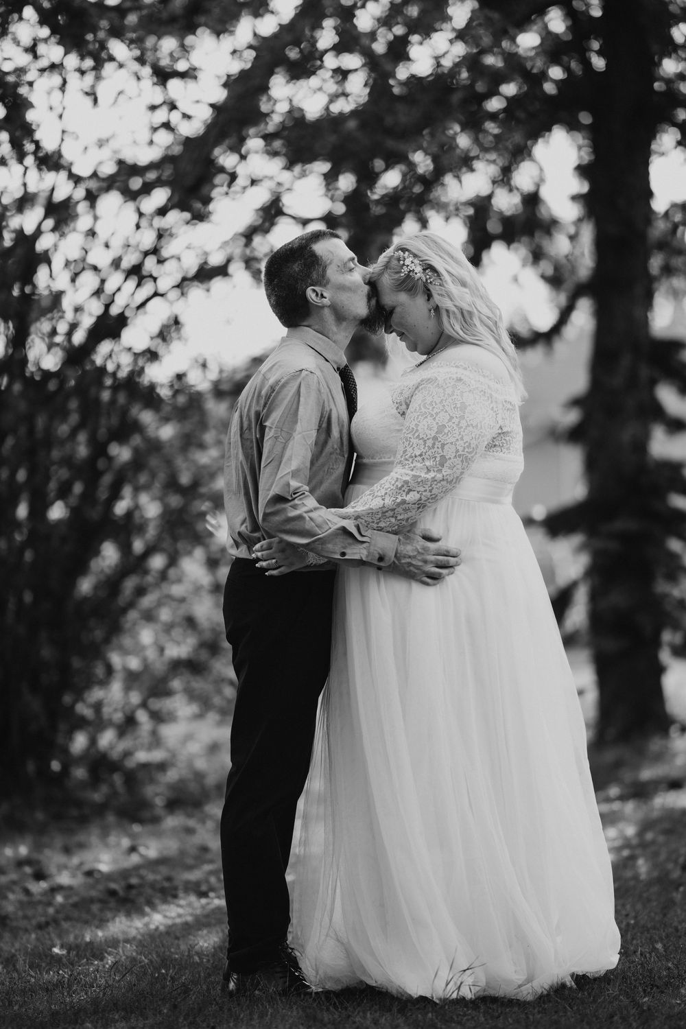 Black and white photo of bride and groom sharing a romantic kiss outdoors under lush green trees.