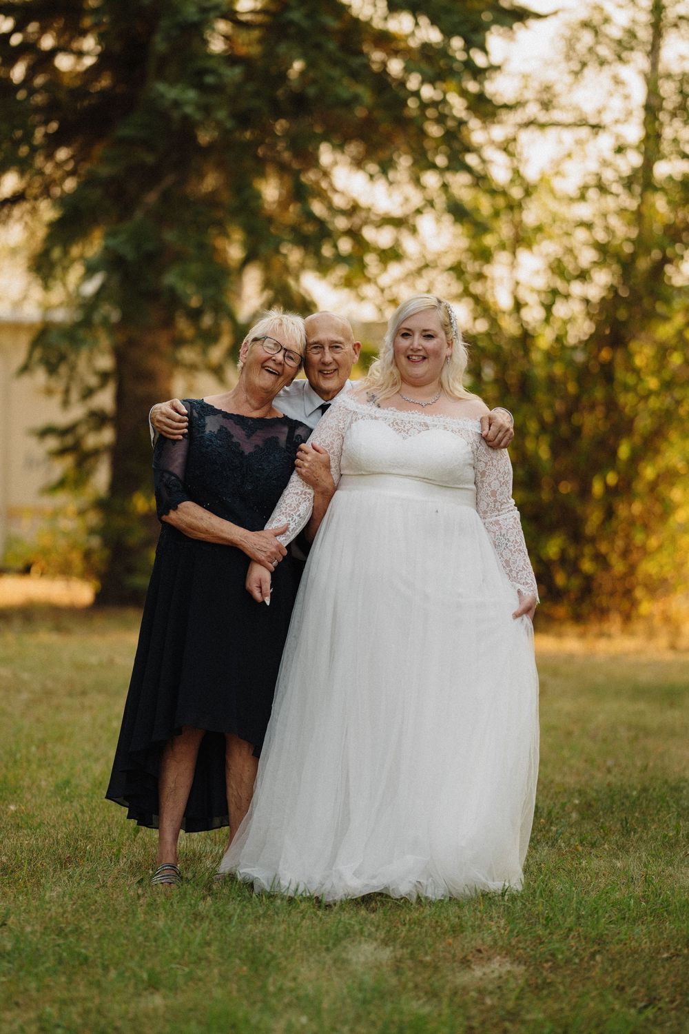 Smiling bride in white lace wedding gown poses with elderly couple in a sunny outdoor garden setting.