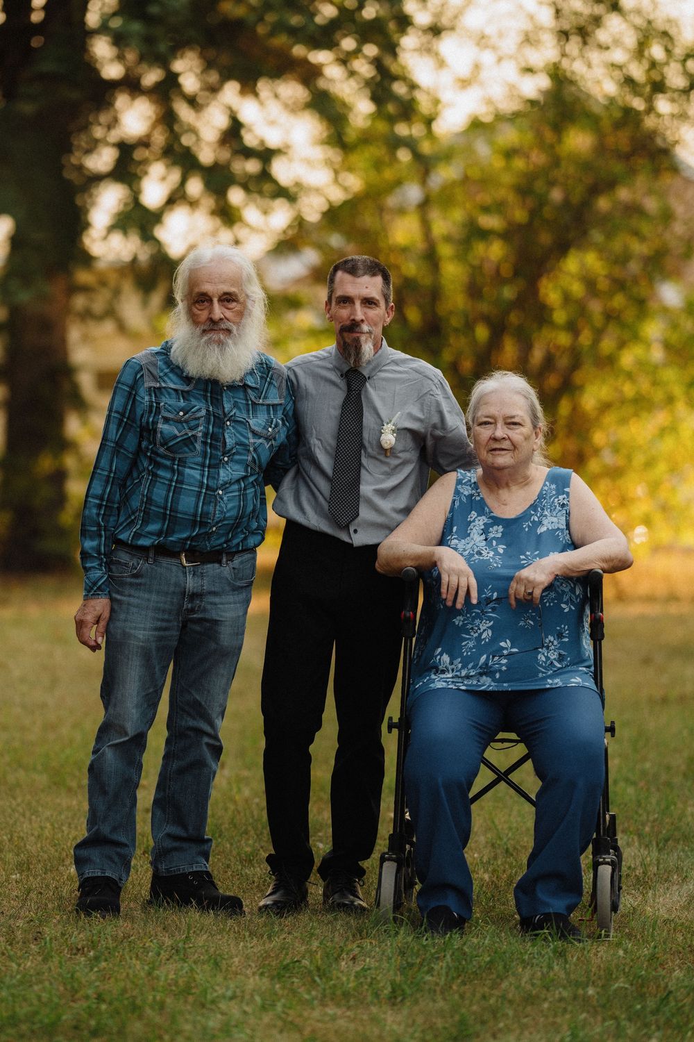 Groom in gray shirt poses outdoors with elderly bearded man and older woman in wheelchair at golden hour.