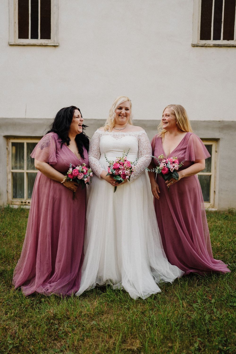 Bride in white lace gown stands between two bridesmaids in dusty rose tulle dresses holding pink bouquets.