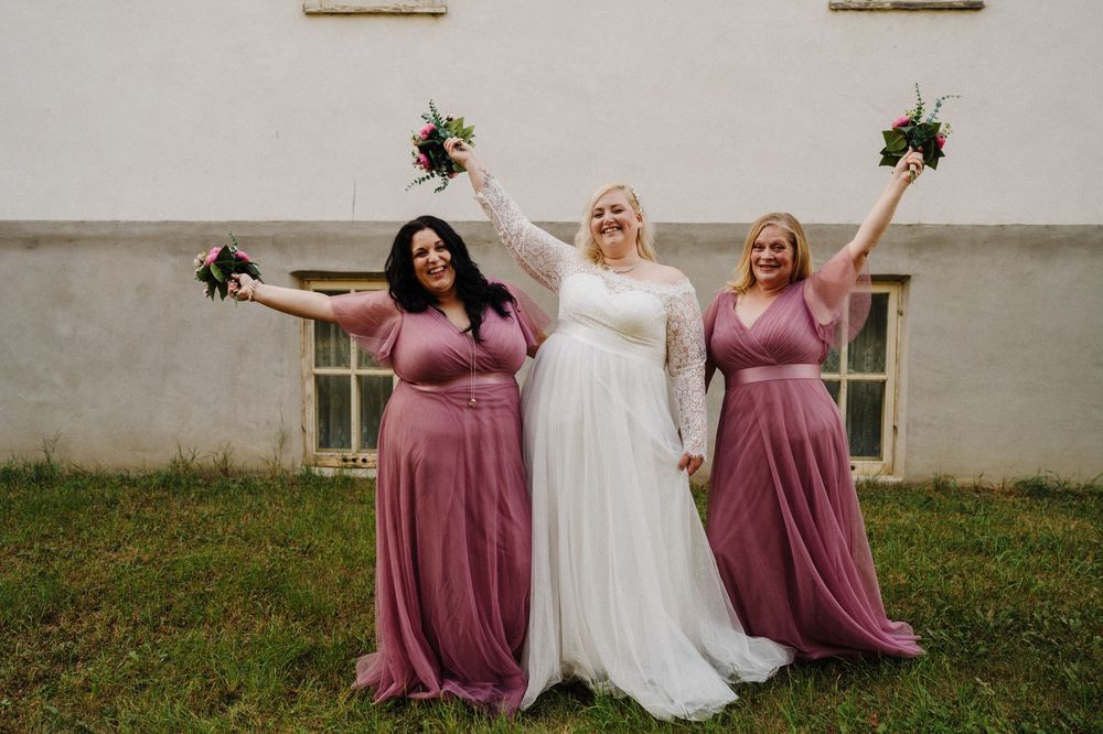 Bride and two bridesmaids in mauve tulle gowns celebrate joyfully with raised bouquets outside a white building.