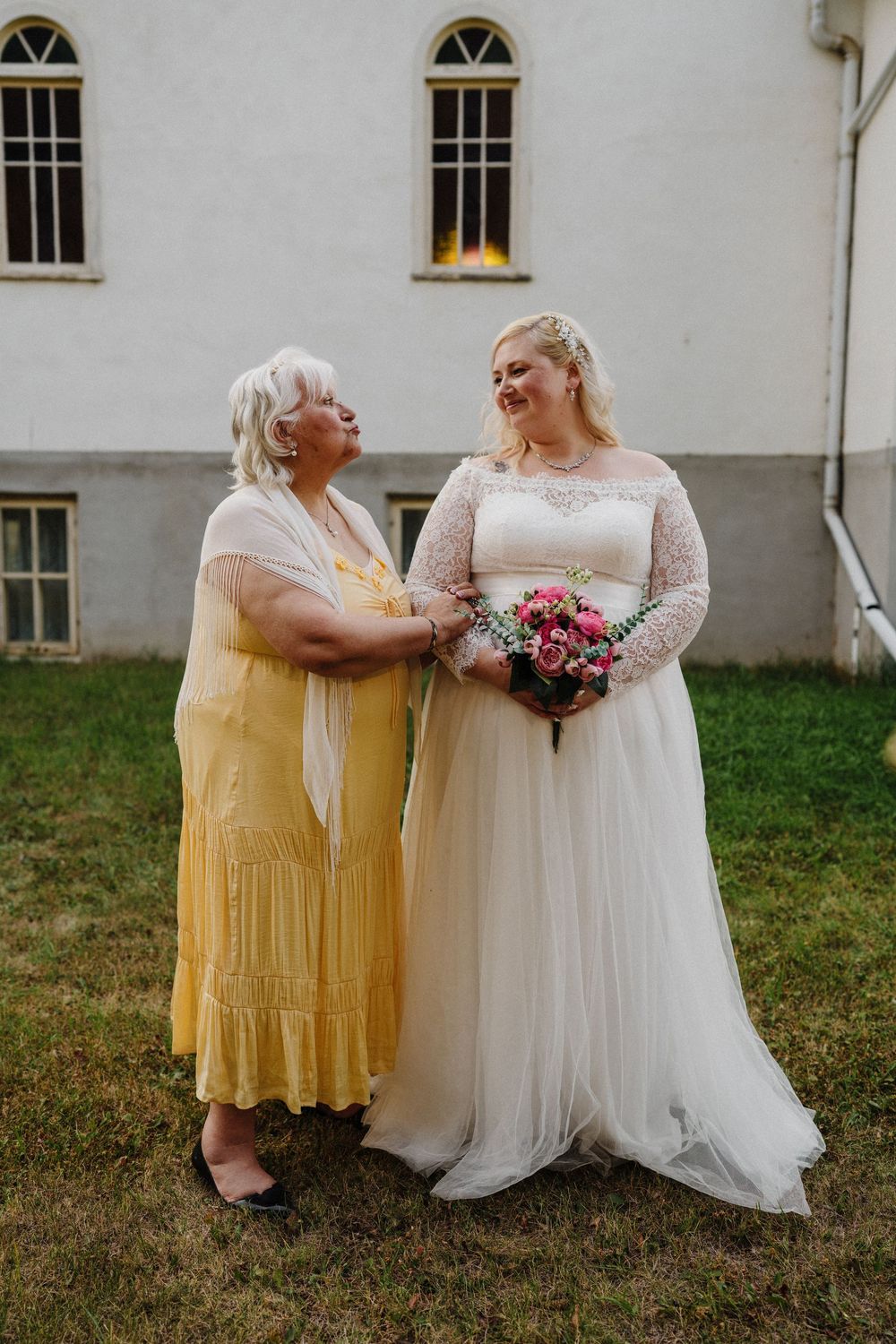 A bride in a white lace gown holds a pink bouquet while sharing a joyful moment with an older woman in a yellow dress.