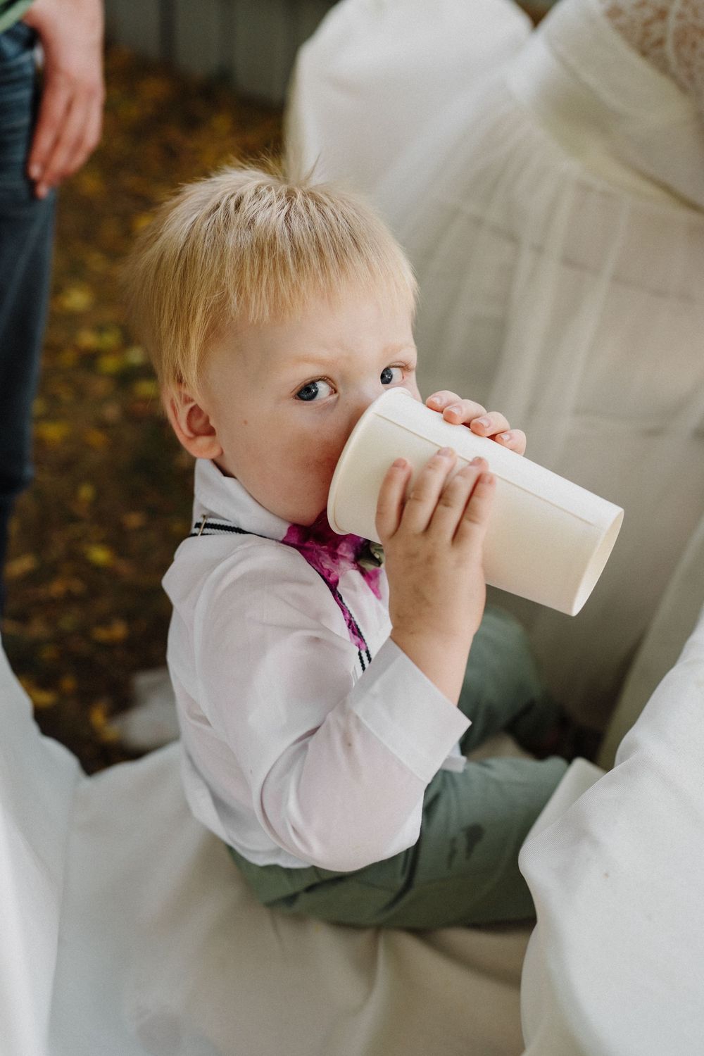 An adorable toddler boy in suspenders and a bow tie drinks from a white cup at a wedding celebration.