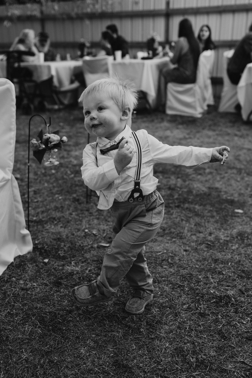 A cute toddler boy in suspenders and bow tie dances joyfully on the grass at an outdoor wedding reception.