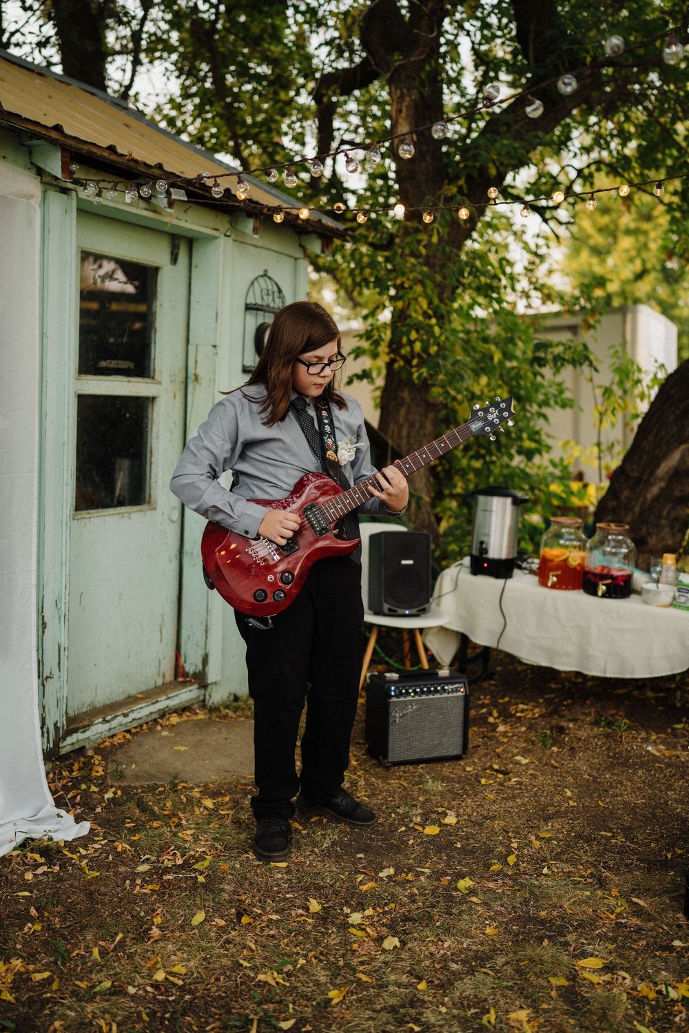 A young guitarist in glasses plays a red electric guitar at an outdoor wedding reception near a vintage shed.