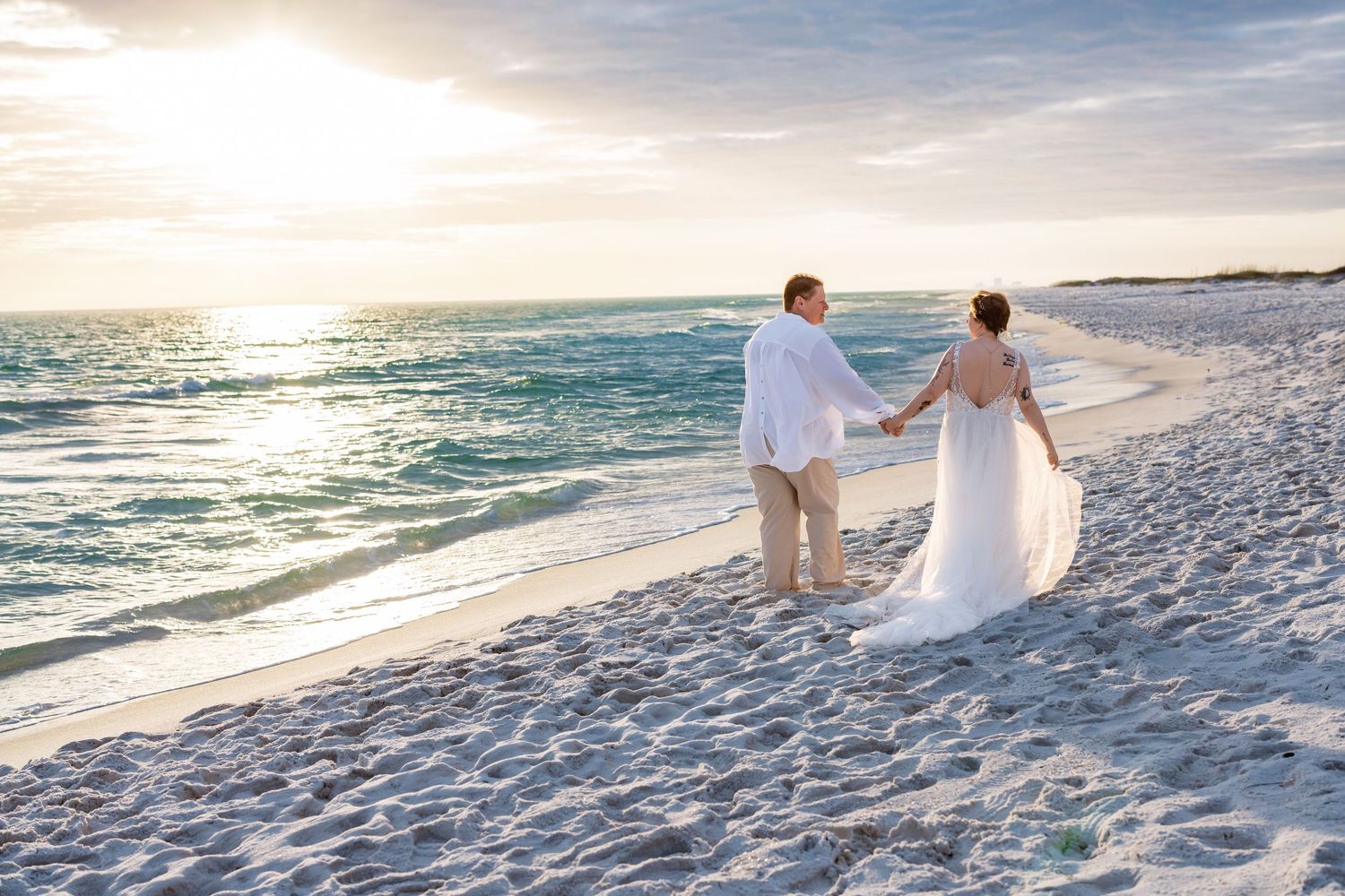 Two people in white attire walk hand in hand along a pristine white sand beach at sunset.