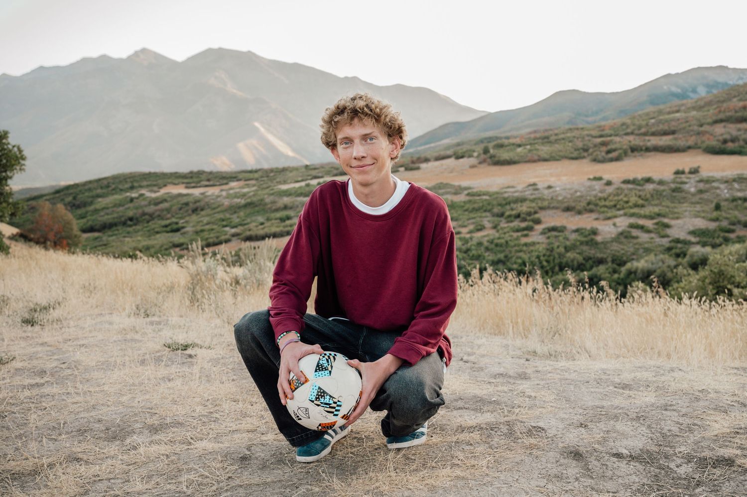 Person holding soccer ball while crouching in mountain field.