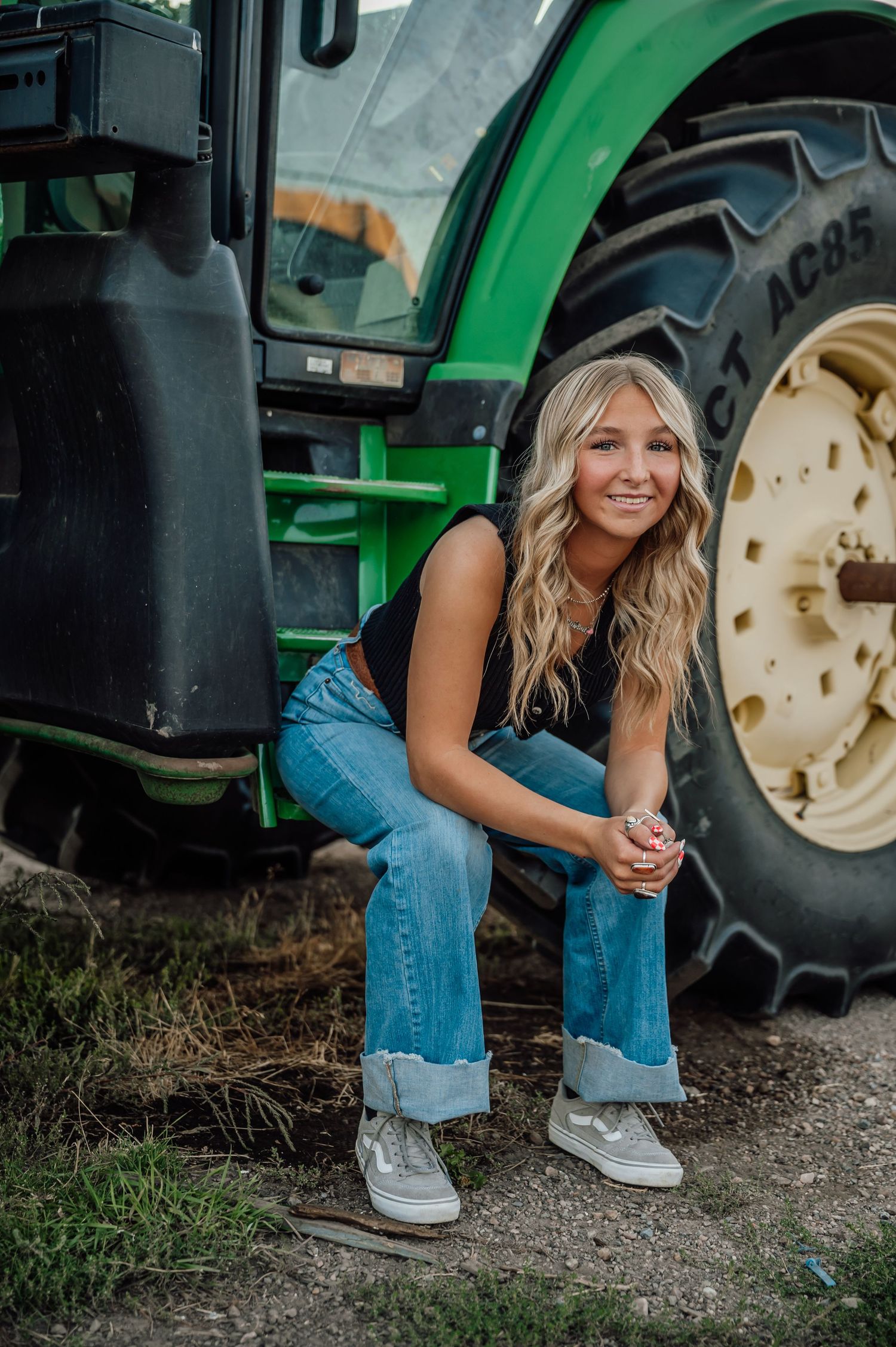 Person in denim sitting by large green tractor wheel in rural setting.