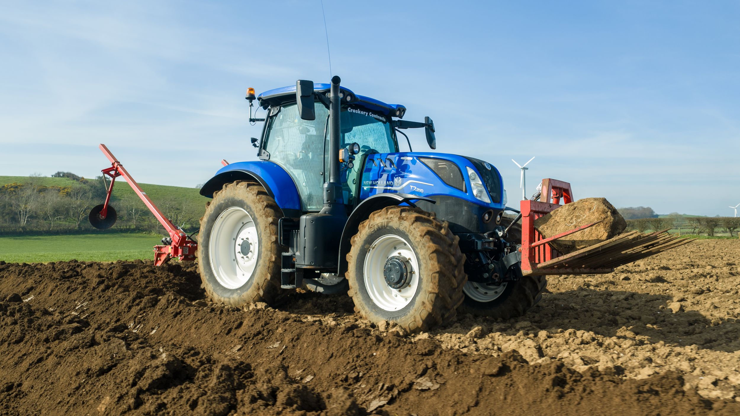 Blue New Holland tractor plowing a field on a sunny day, turning rich dark soil in a rural agricultural landscape.