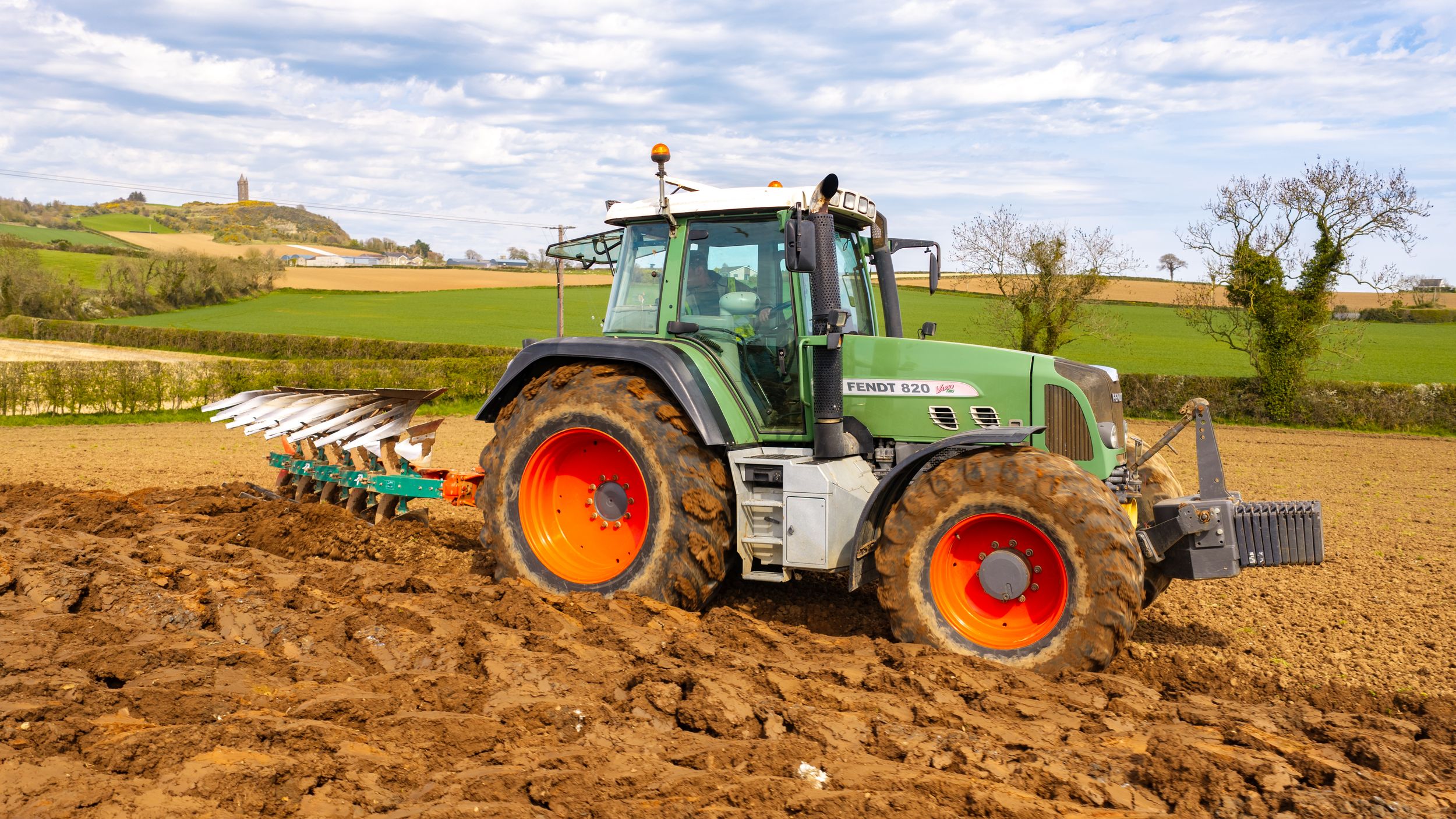 Green Fendt tractor plowing agricultural field with multi-furrow plow attachment on sunny day in rural countryside.