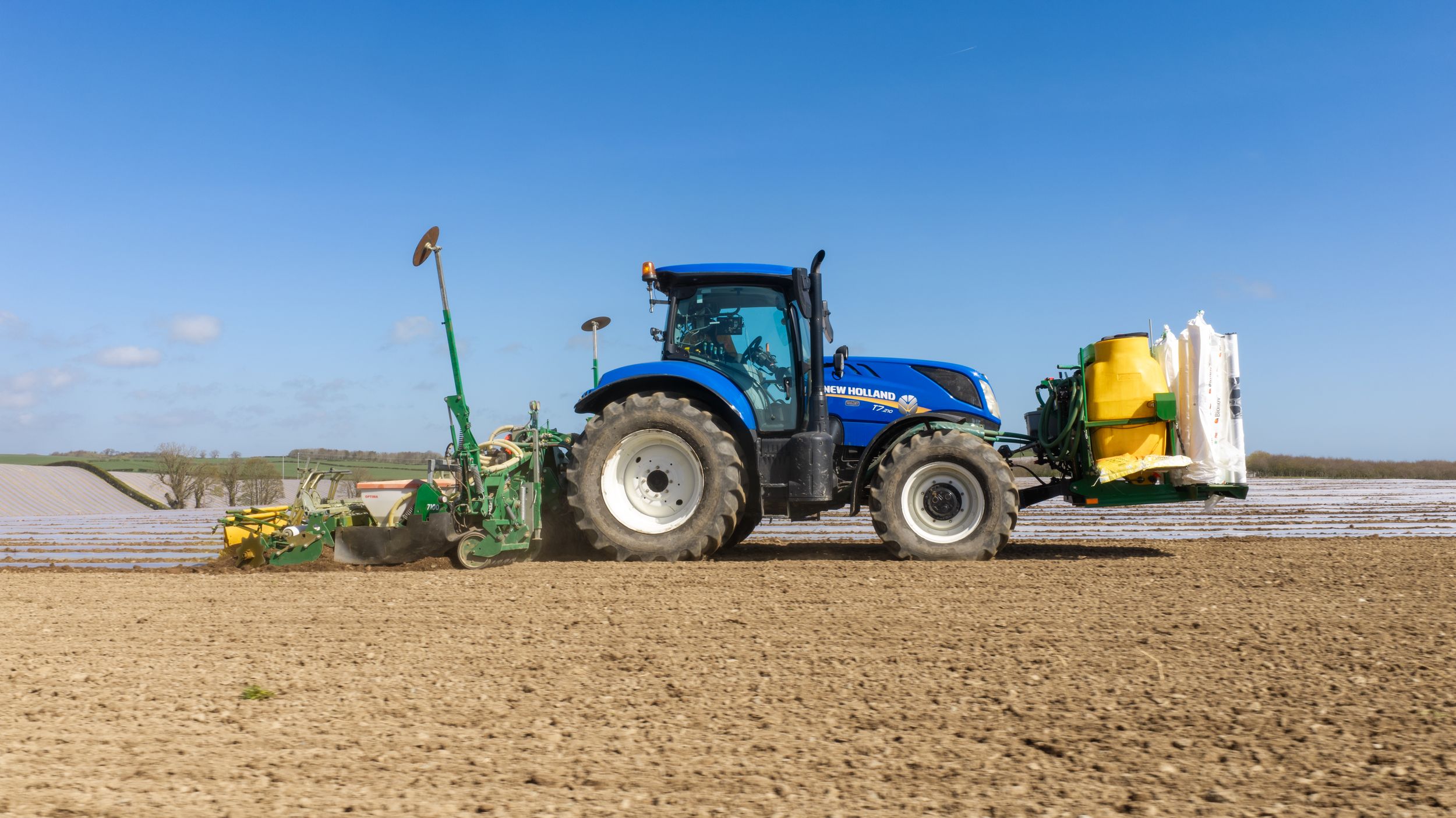 Blue New Holland tractor with green seeder attachment working in a plowed agricultural field under a clear blue sky.