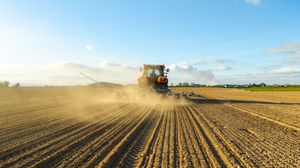 Red tractor plowing dusty agricultural field at sunset, creating large dust clouds across vast farmland under blue sky.