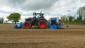 A black Fendt tractor with red wheels tilling a large field with blue agricultural equipment attached.
