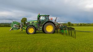 John Deere tractor with hay rake attachment working in a green field under cloudy skies.