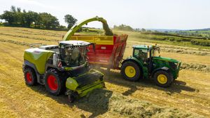 A Claas forage harvester transfers cut grass into a John Deere tractor trailer during hay harvesting in a sunny field.
