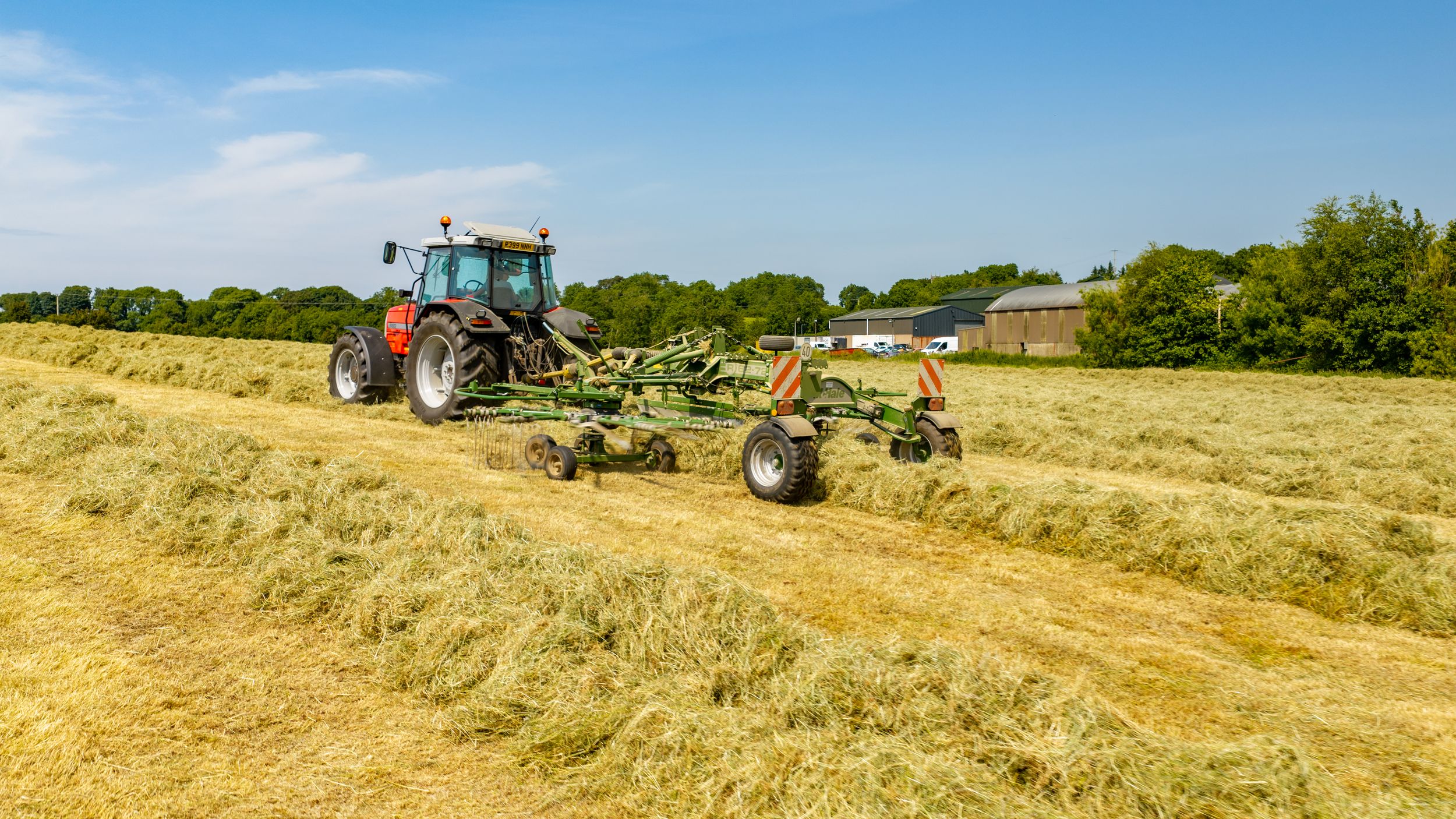 Red tractor with green hay rake tedding cut grass in a sunny farm field during summer harvest season.