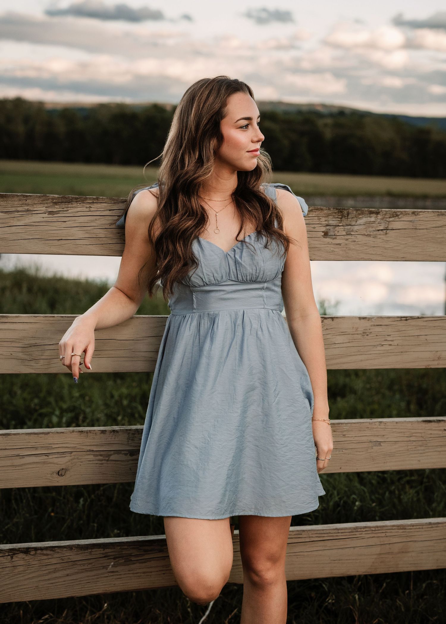 Woman in blue dress poses against wooden railing at sunset.