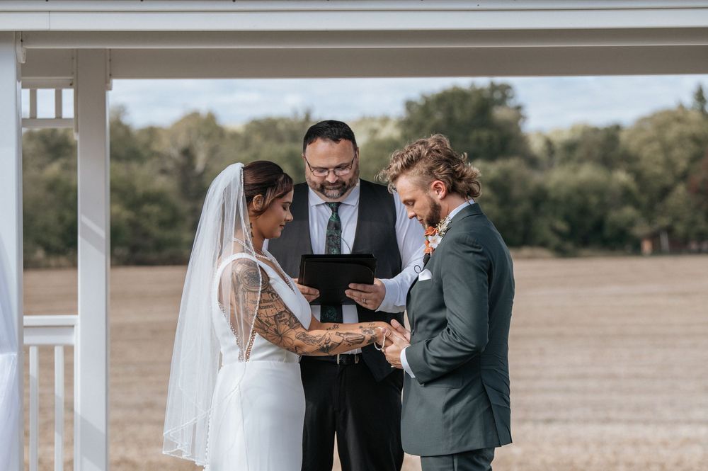 An intimate outdoor wedding ceremony taking place under a white gazebo with guests seated on white chairs.
