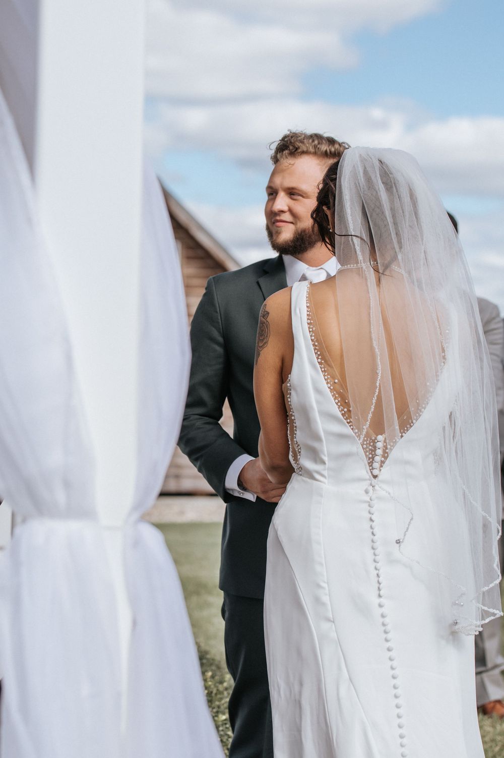 A bride in an elegant white dress with button details and flowing veil during an outdoor ceremony.