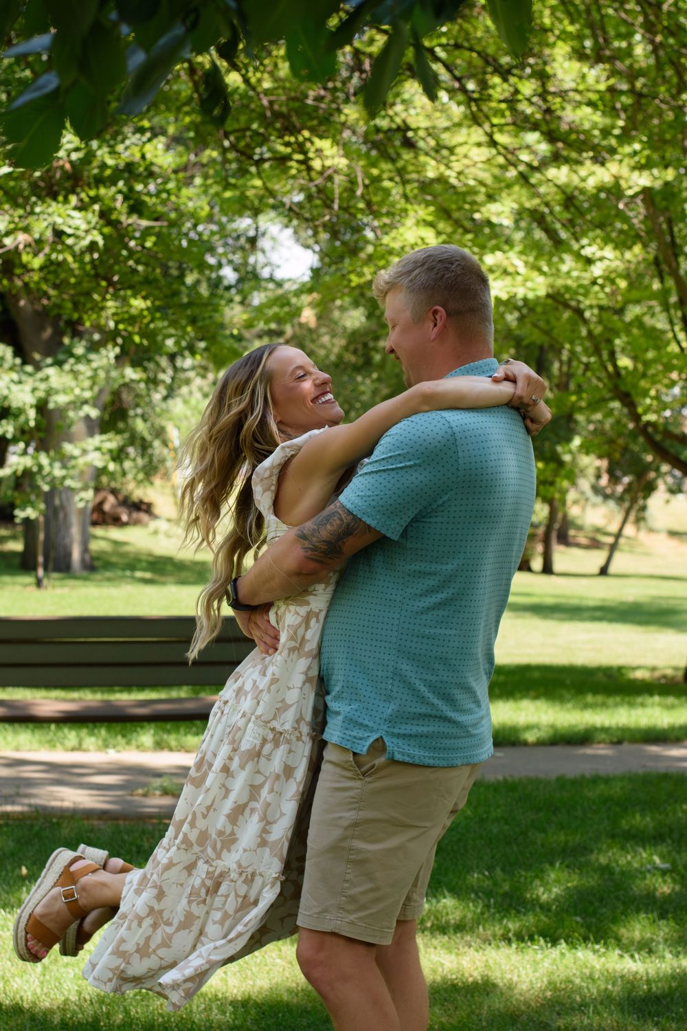 Playful couple dancing and spinning in a sunlit park with lush green trees.