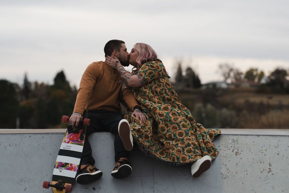 Couple sitting casually on concrete wall with water and cloudy sky in background.