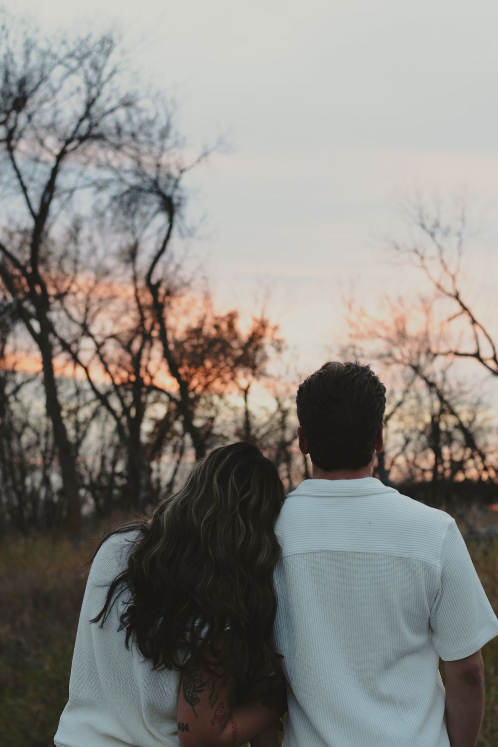 Silhouetted couple in white clothing stands together at sunset with bare trees in background.