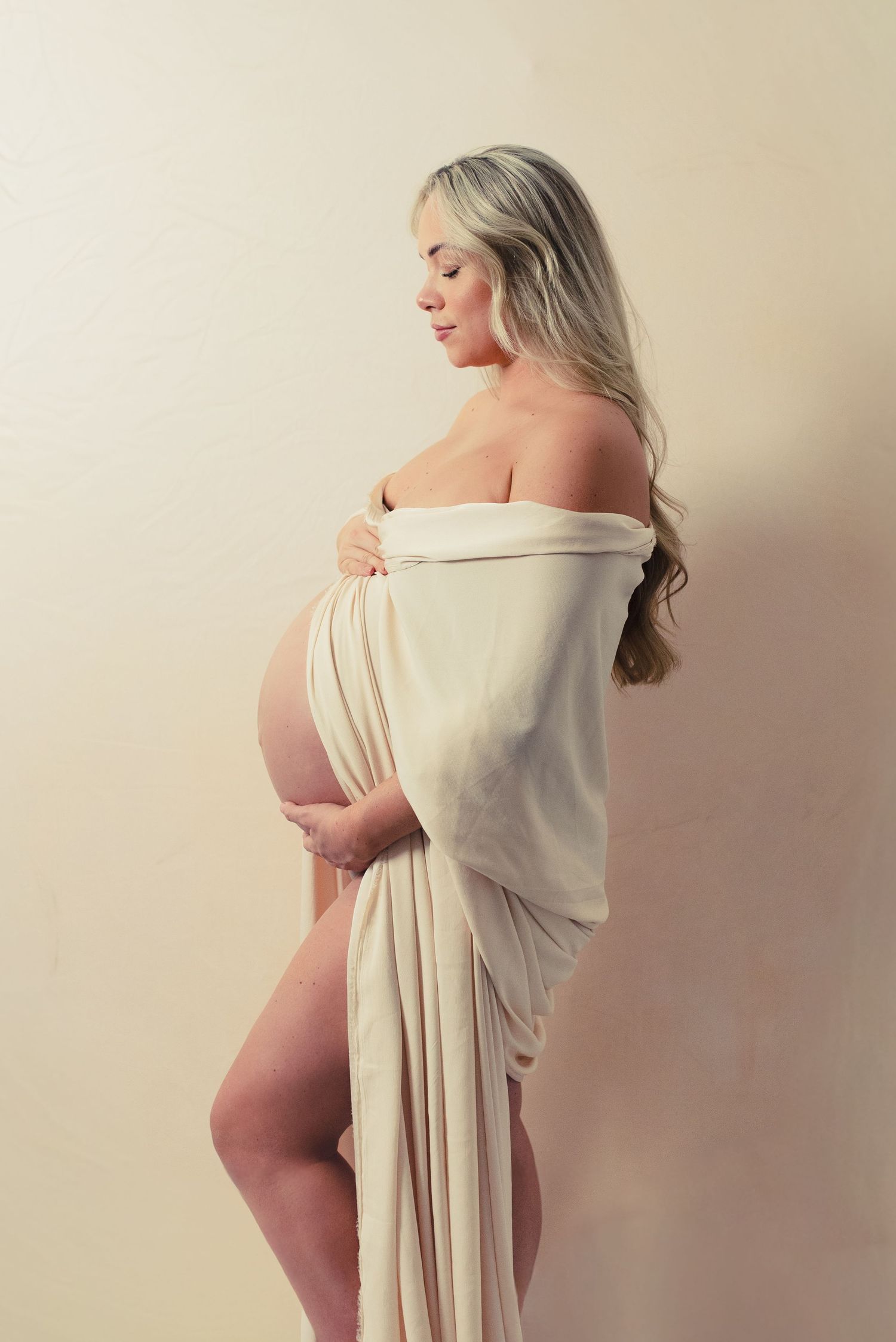 Artistic maternity photo series showing silhouette of pregnant woman draped in flowing cream fabric against neutral backdrop.