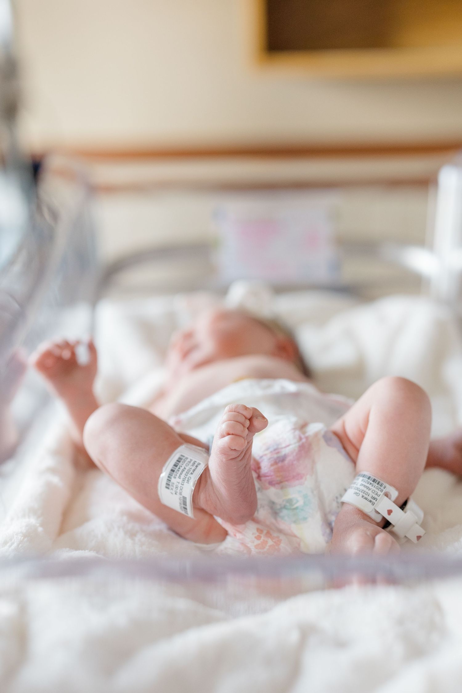 A newborn baby's tiny feet with hospital identification bands rest on soft white blankets.
