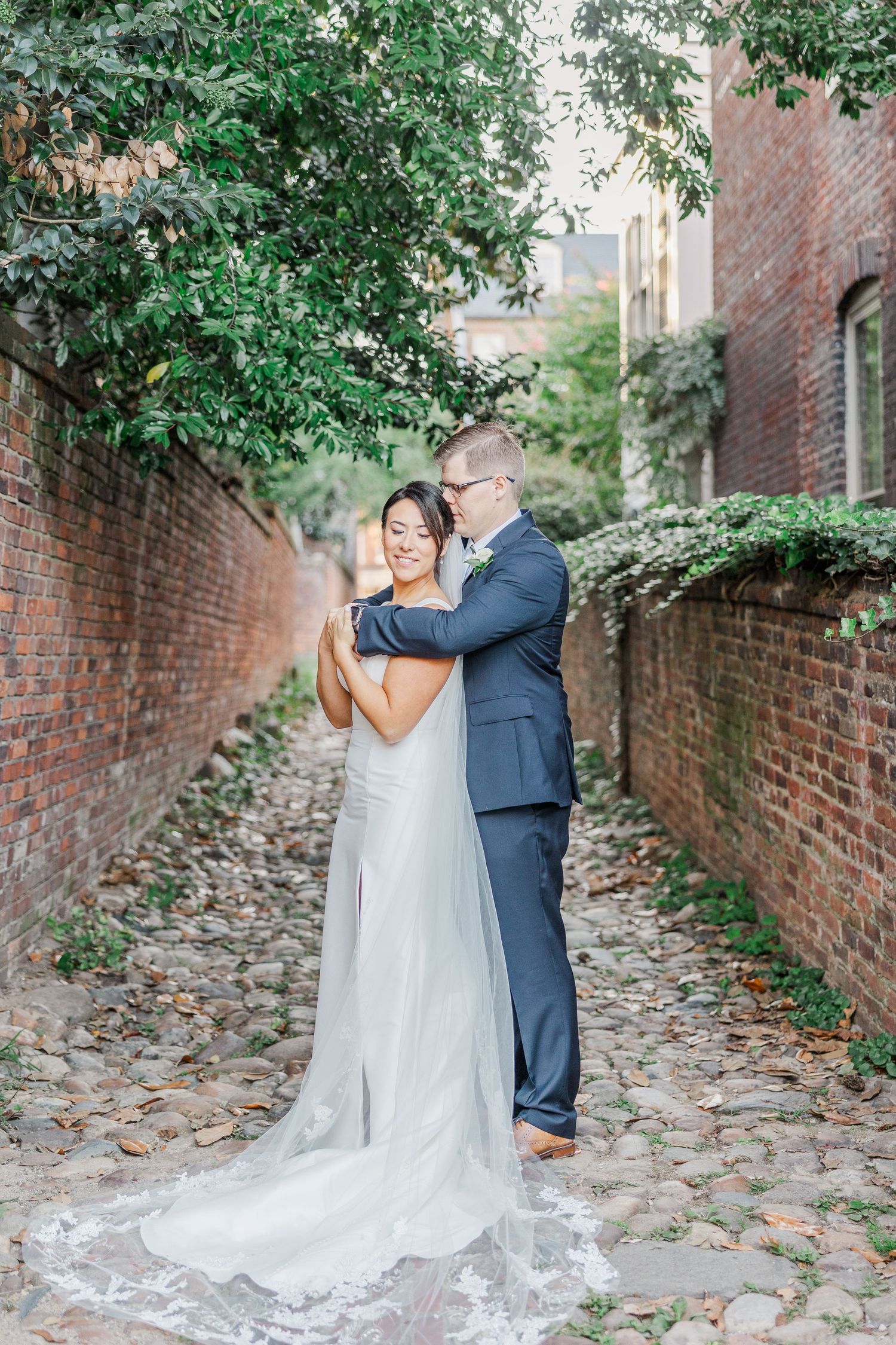 A couple dances together on a cobblestone path surrounded by brick walls and overhanging greenery.