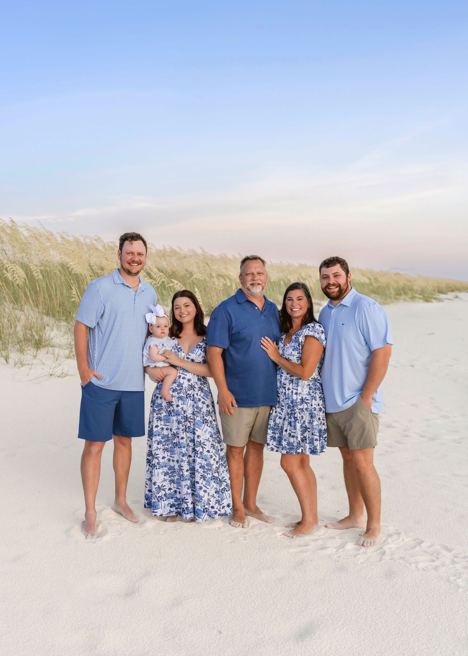 Family of six posing on white sand beach in coordinating blue outfits, smiling together near coastal dunes.