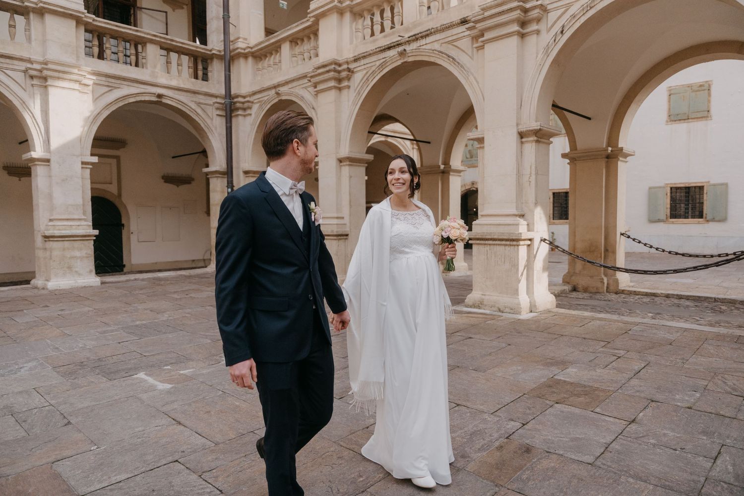 A couple walks through an elegant stone courtyard with arched columns and classical architecture.