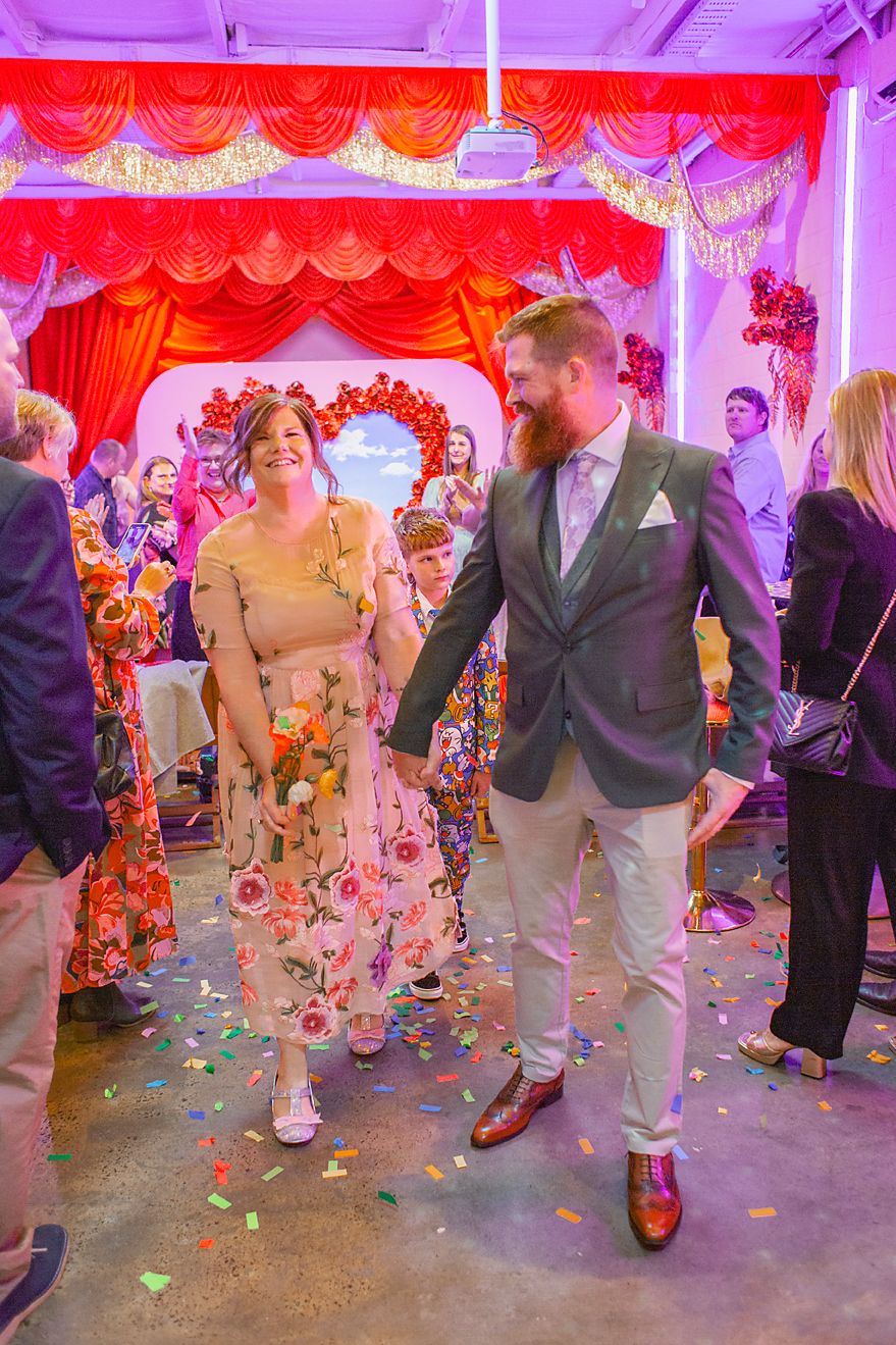 Feet of wedding guests dancing on a confetti-covered floor under purple and red lighting at a reception celebration.