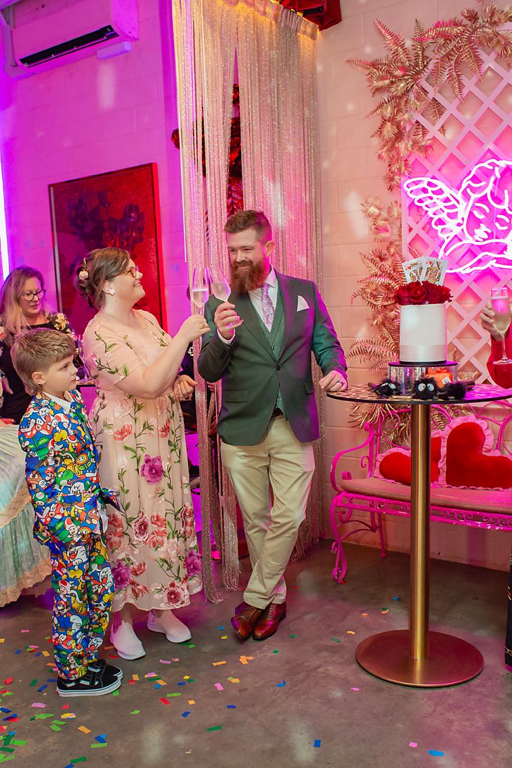People dancing and celebrating in a pink-lit room with floral decorations and confetti on the floor.