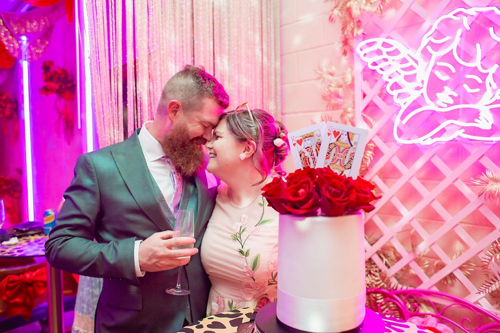 A wedding couple shares intimate moments near their white tiered cake in a pink-lit reception venue.