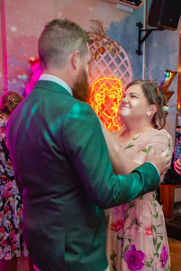 A couple dancing together at a party with colorful neon lighting and floral decorations in the background.