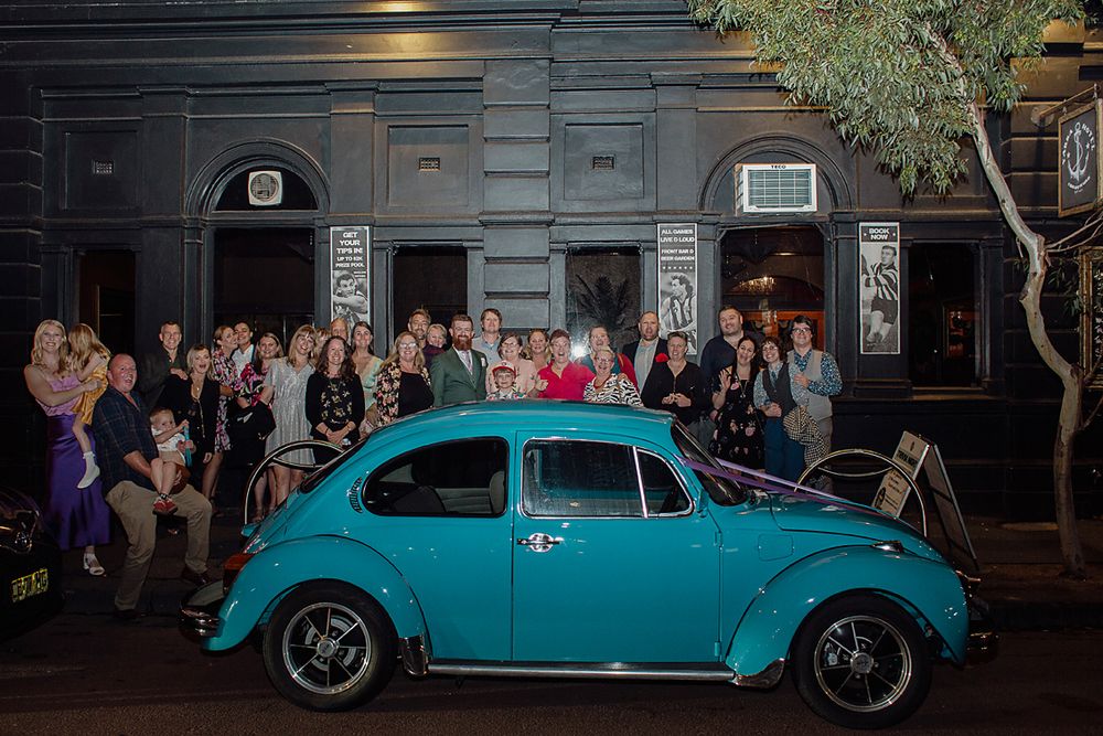 A bright turquoise vintage Volkswagen Beetle parked at night outside a venue with a crowd gathered behind it.