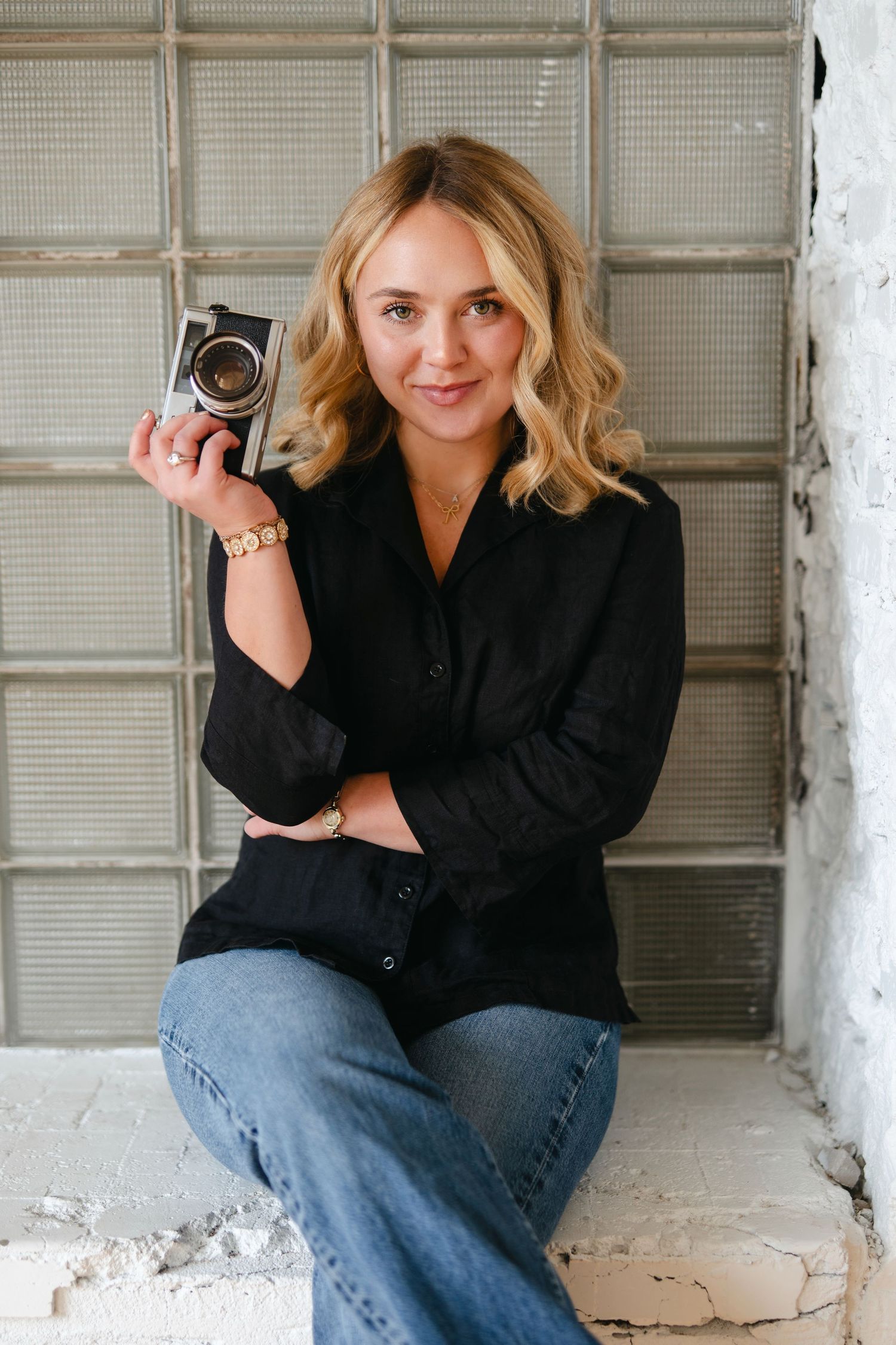 A person in a black blazer and jeans sits casually against a white wall with glass block windows.