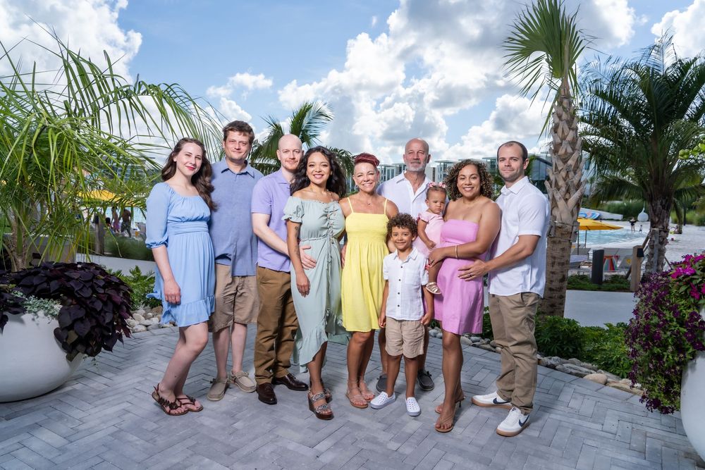 A happy family group poses together outdoors against a tropical backdrop with palm trees and blue sky.