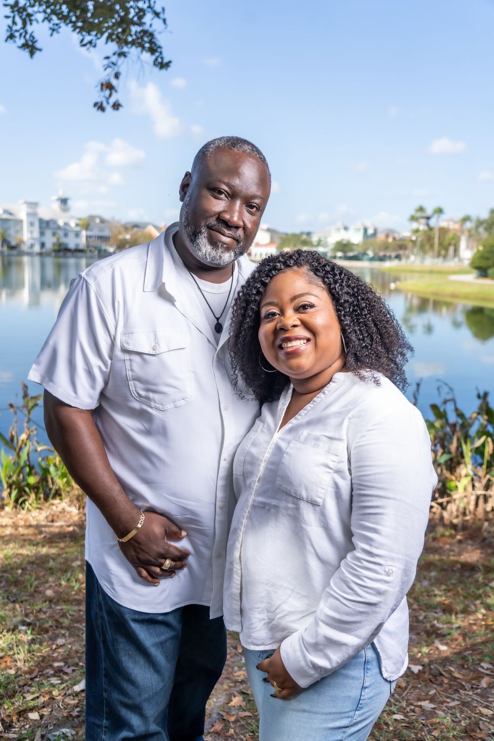 A couple wearing white shirts smiles together while standing by a scenic lake on a sunny day.