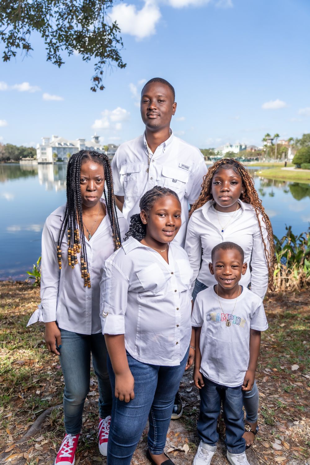Family wearing coordinated white shirts and jeans pose together outdoors near a lake on a sunny day.