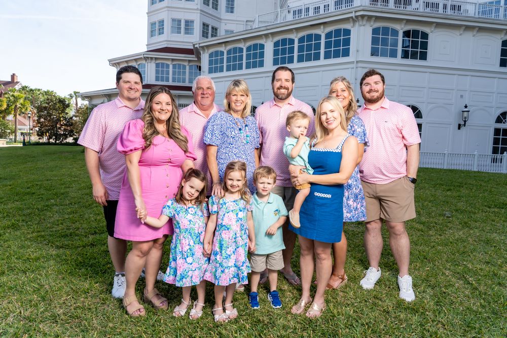 A group poses together on a lawn in front of a white building, wearing pink and blue summer attire.