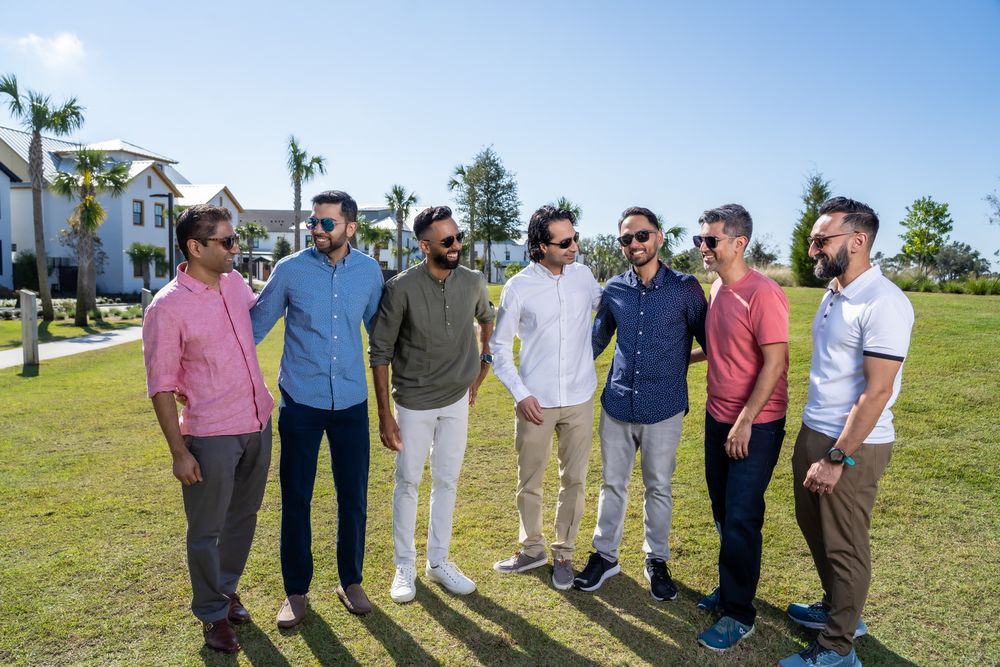 Group of friends in casual attire standing together on green grass against a bright blue sky background.