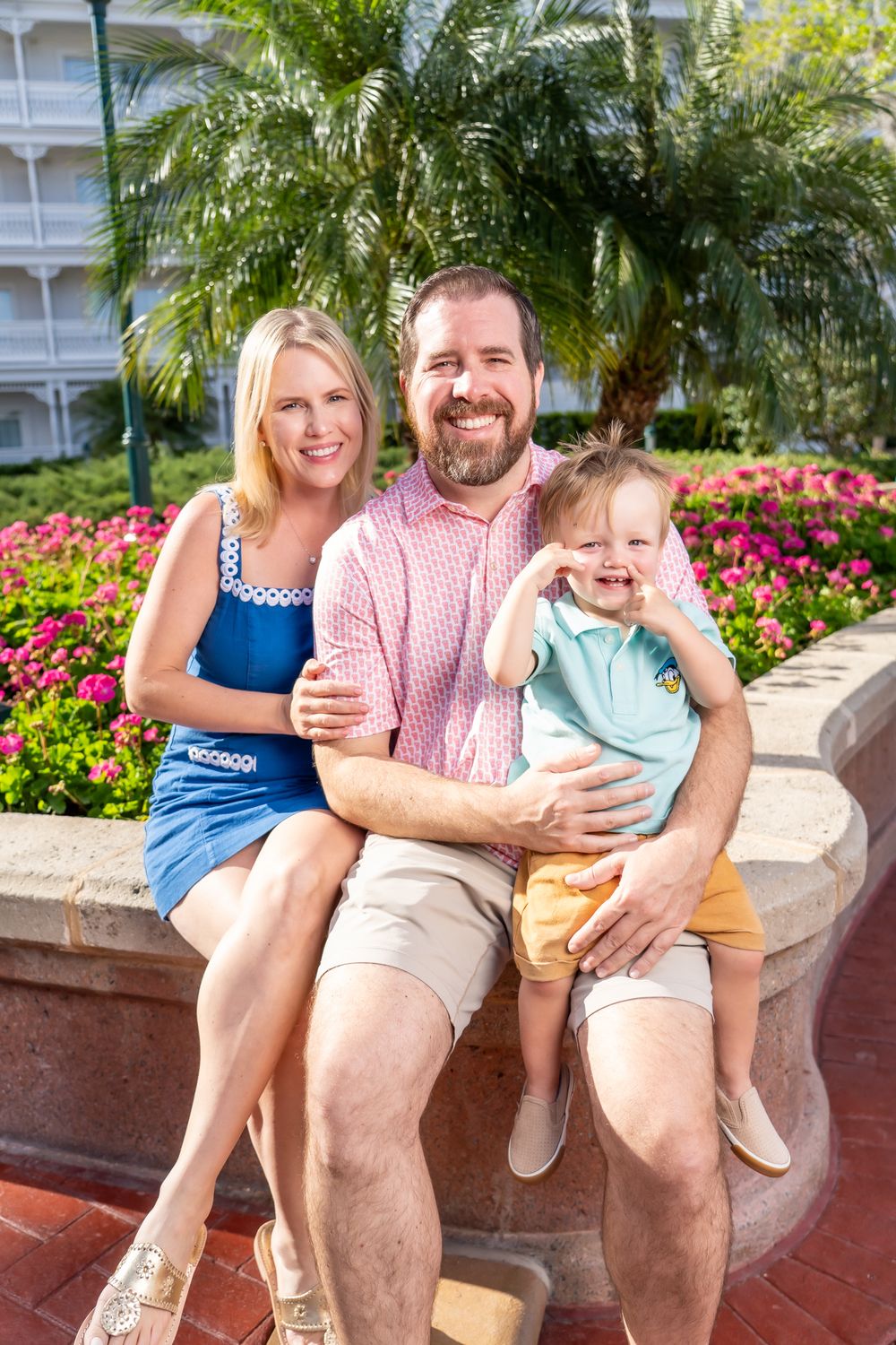 A family sits together on a stone wall surrounded by vibrant pink flowers and tropical palm trees at a resort.