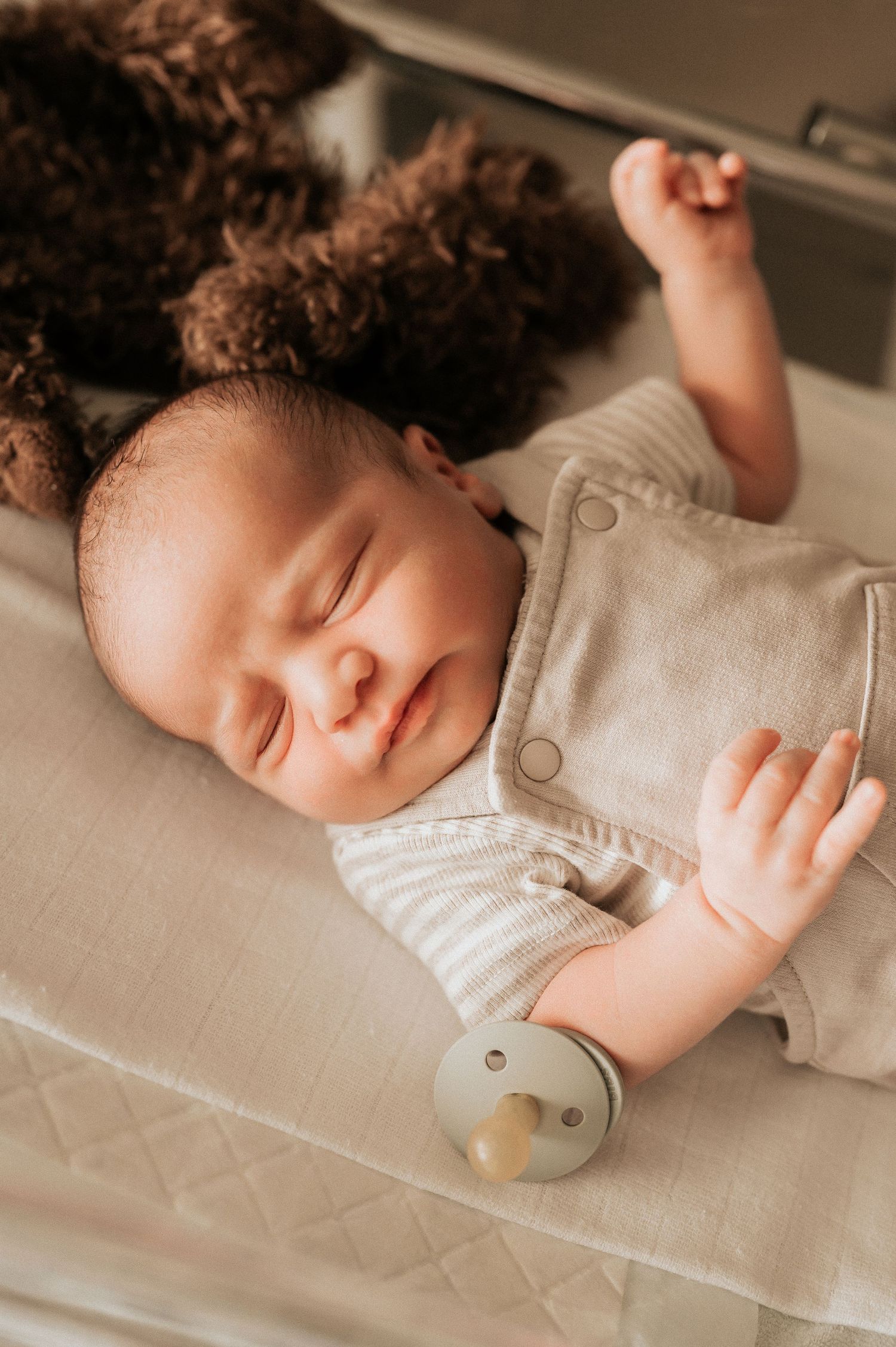 Slapende baby in een beige outfit rust rustig op een lichte ondergrond met een fopspeen in de buurt, vastgelegd in West-Vlaanderen.