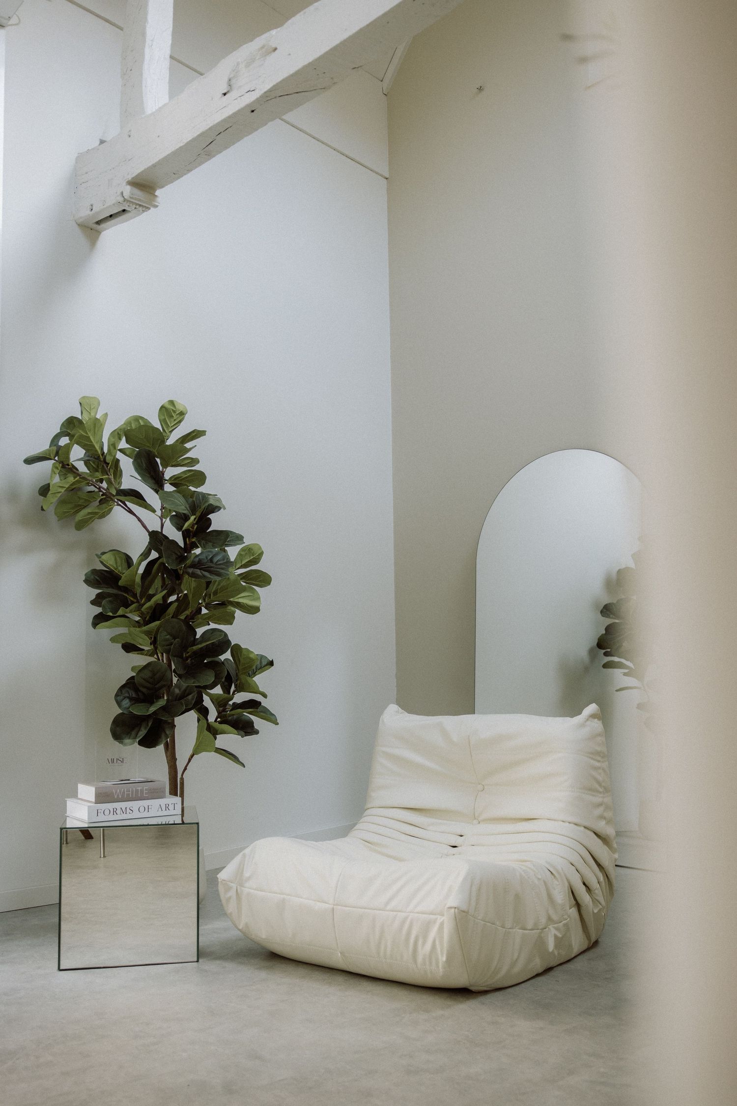 Minimalist white interior with a plush Togo chair, mirrored side table, and fiddle leaf fig plant against white walls.
