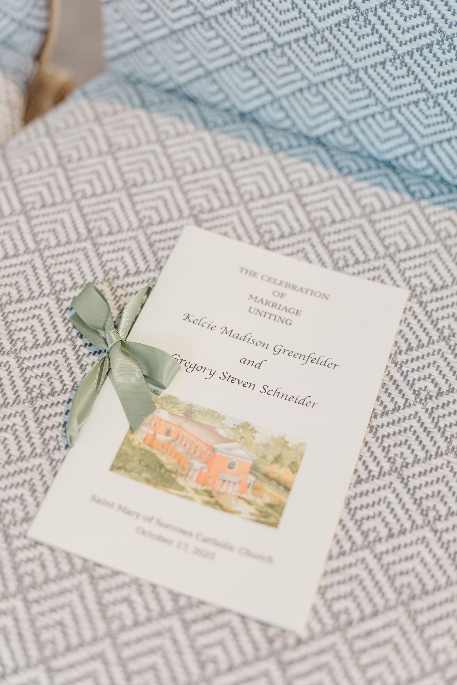 Wedding invitation cards with watercolor illustration of a coral-colored house and sage green ribbon details on textured fabric.