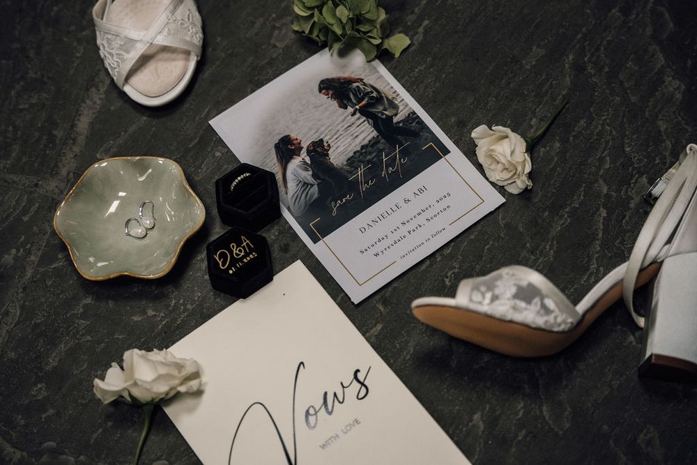 Wedding details flatlay with vows, photograph, jewelry and white shoes on dark surface.