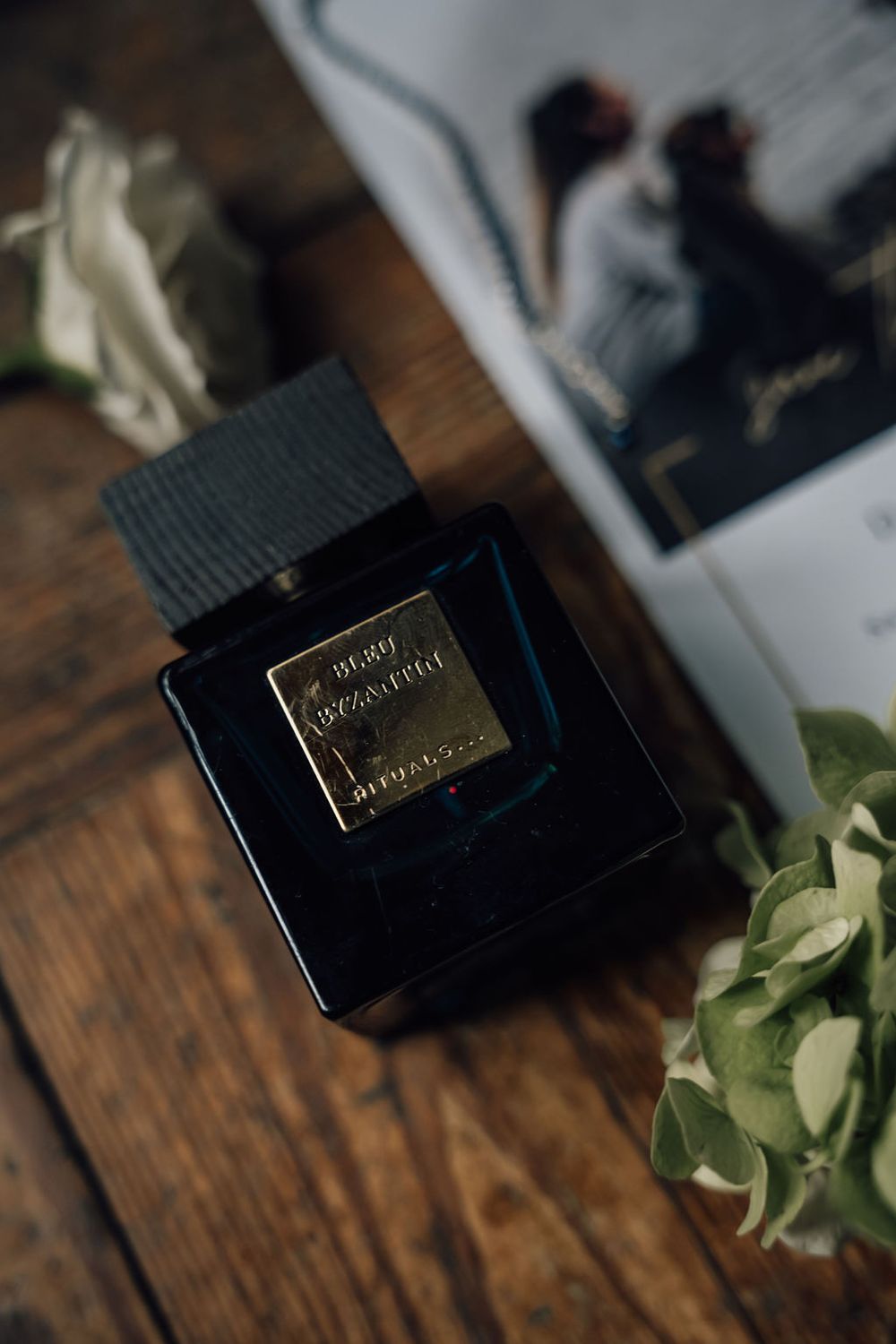 Elegant black perfume bottle with gold label next to floral elements and photograph.
